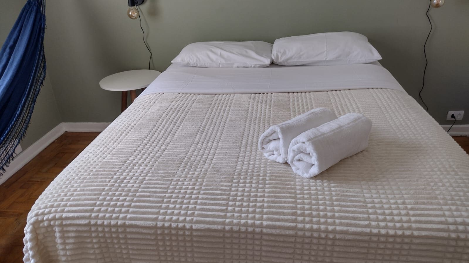 A neatly made bed with a textured white duvet, a rolled white towel on the bed, and a round white nightstand beside it.