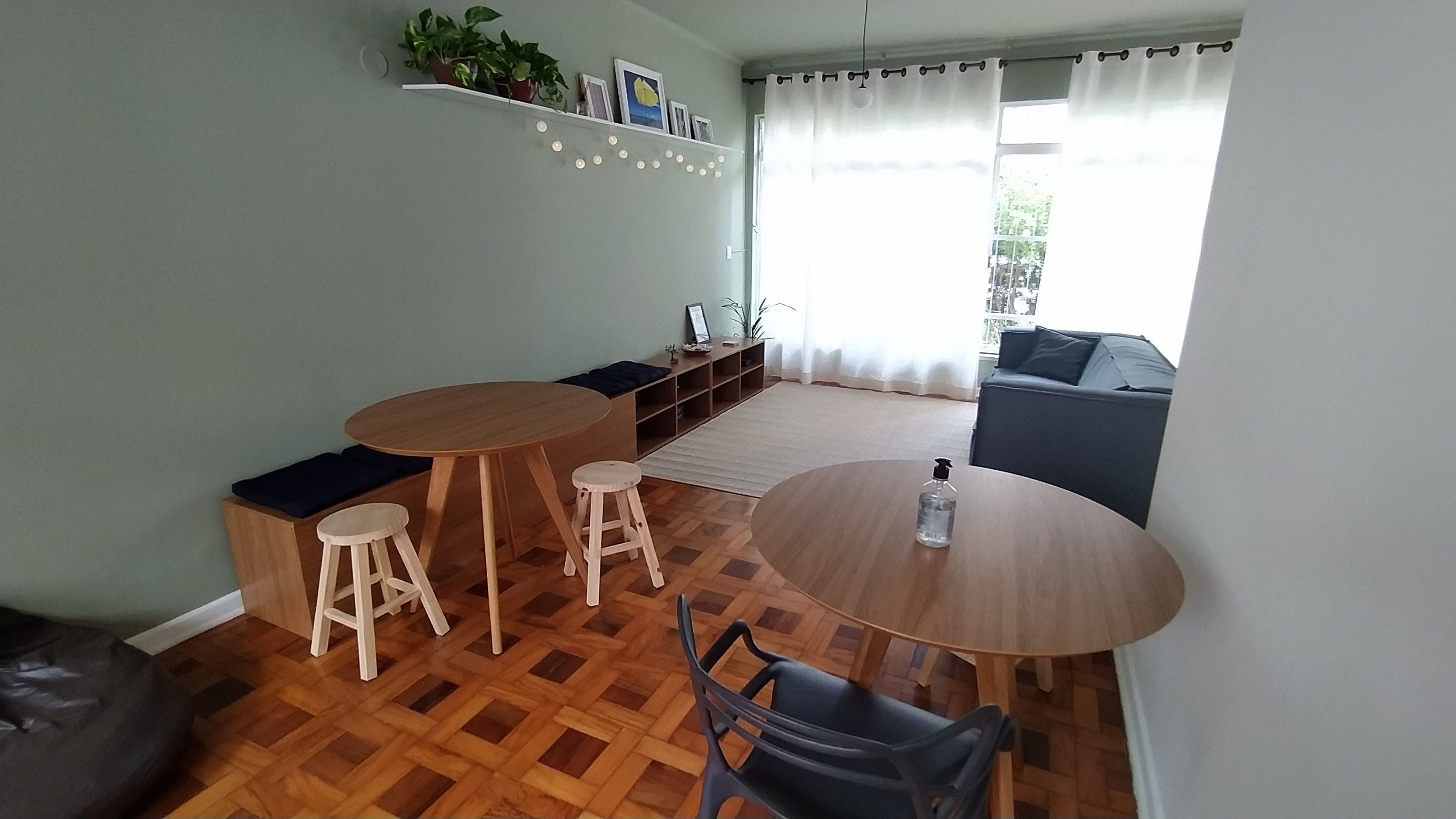Ein Essbereich mit einem runden Tisch, zwei Hocker und einem Stuhl. Im Hintergrund befindet sich eine Couch und ein Fenster mit Vorhängen.