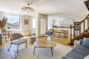 Un espacio habitable amplio con piso de madera, una mesa de centro redonda y un sofá. Se ve una cocina al fondo con gabinetes blancos y una escalera de madera.