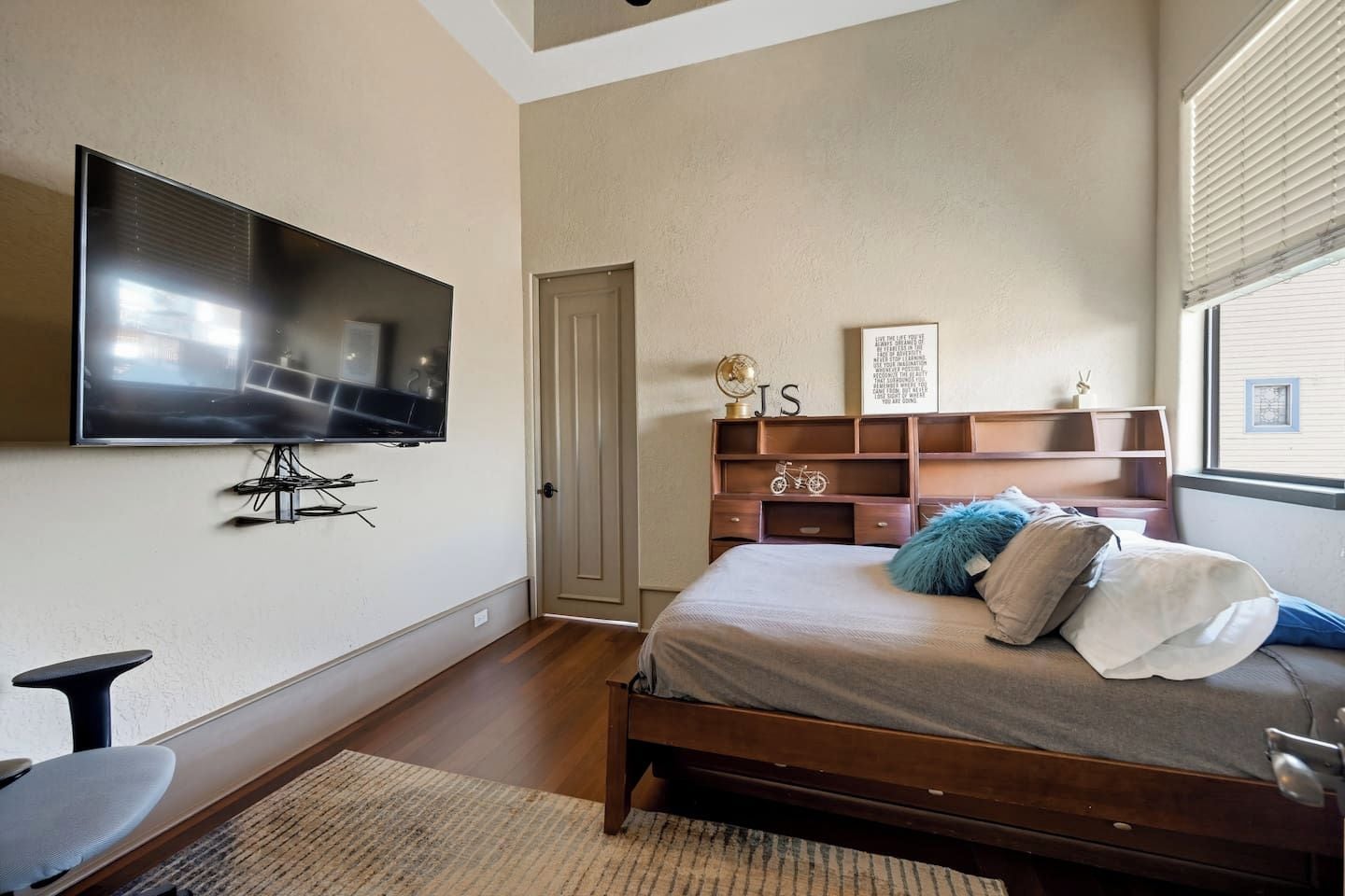Un dormitorio con cama, televisor montado en la pared, cabecera de madera, una puerta y una ventana con persianas.