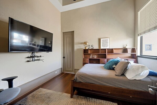 Un dormitorio con cama, televisor montado en la pared, cabecera de madera, una puerta y una ventana con persianas.