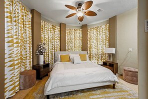 A bedroom with a bed, nightstands, a lamp, a ceiling fan, and patterned curtains.