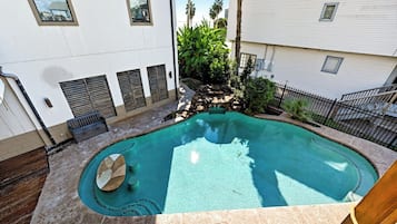 Una piscina con una cascada de piedra, rodeada por un patio con mobiliario y un pequeño jardín.