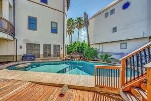 A swimming pool with a wooden deck and a stone border, surrounded by a white building with multiple windows and a blue fence.