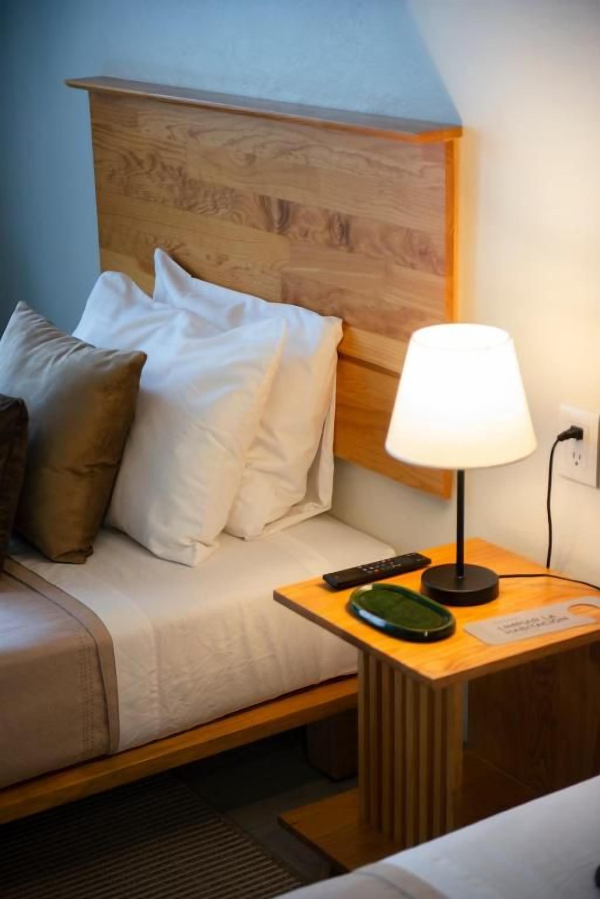 A hotel room with a wooden headboard, a bed with white and brown pillows, a bedside table with a lamp, a remote control, and a nameplate.