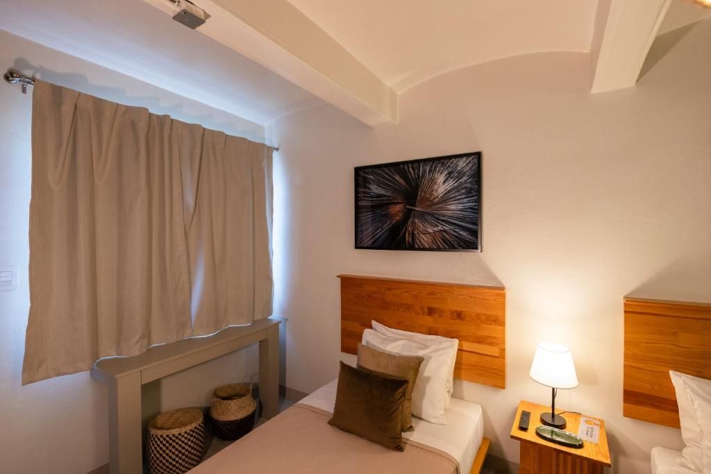 A hotel room with a bed, a headboard, a bedside table with a lamp, a wall-mounted artwork, and a window with curtains.
