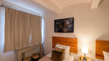A hotel room with a bed, a headboard, a bedside table with a lamp, a wall-mounted artwork, and a window with curtains.