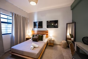 A hotel room with a bed, bedside lamps, a mirror, a small round ottoman, and a bench. - Origen by ÓOL Hotels (Oaxaca)