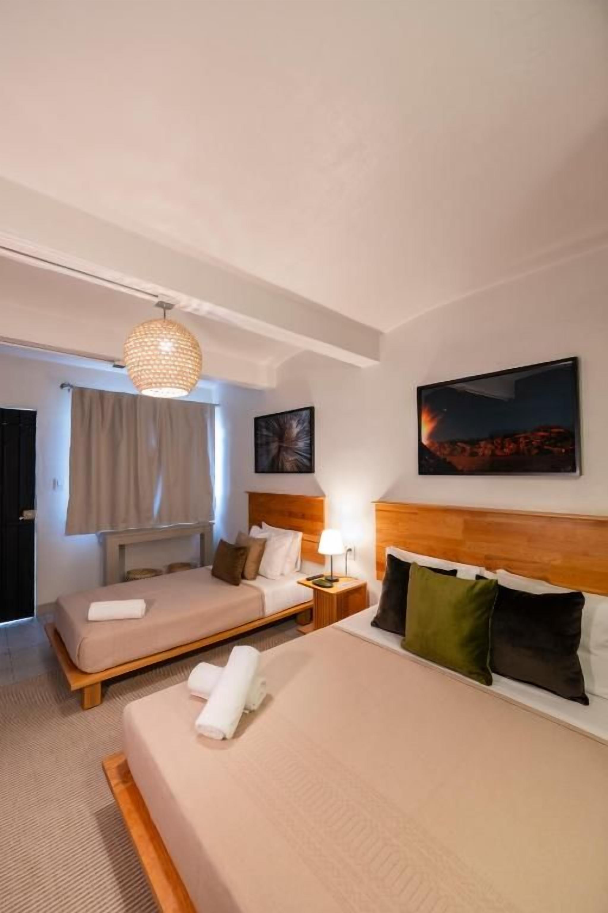 Une chambre d’hôtel avec deux lits, une table de chevet avec une lampe, un grand tableau au mur et une fenêtre avec des rideaux.