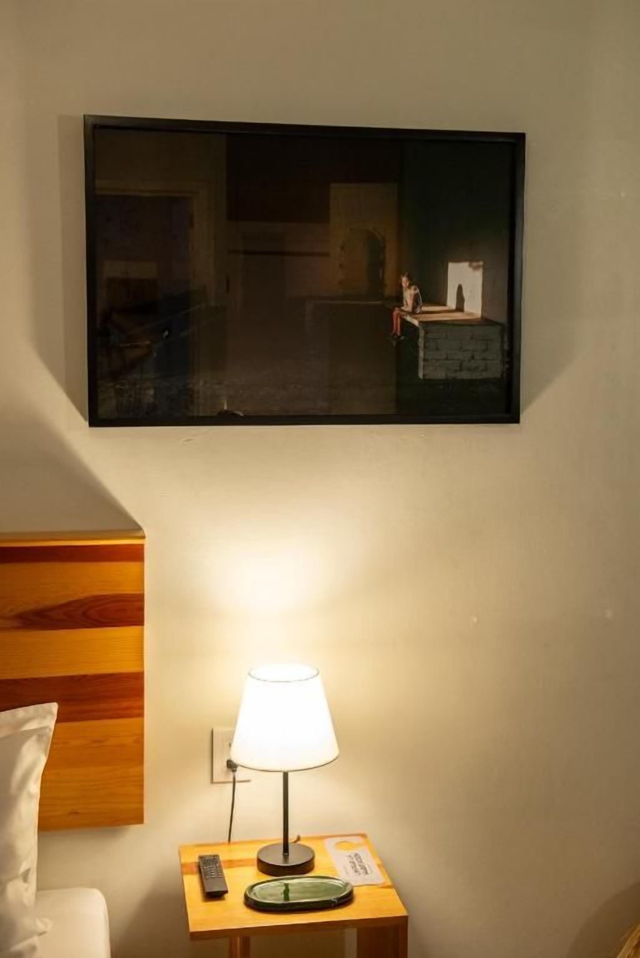 Una habitación de hotel con una cama, una mesita de noche con una lámpara, un control remoto y una obra de arte enmarcada en la pared.