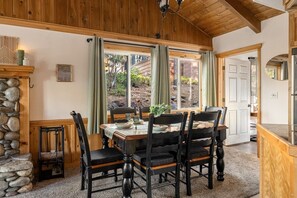 A dining area with a wooden table and chairs, a stone wall, and a view through large windows. - The Village Villa - Hot Tub, Walk to village (Big Bear Lake)