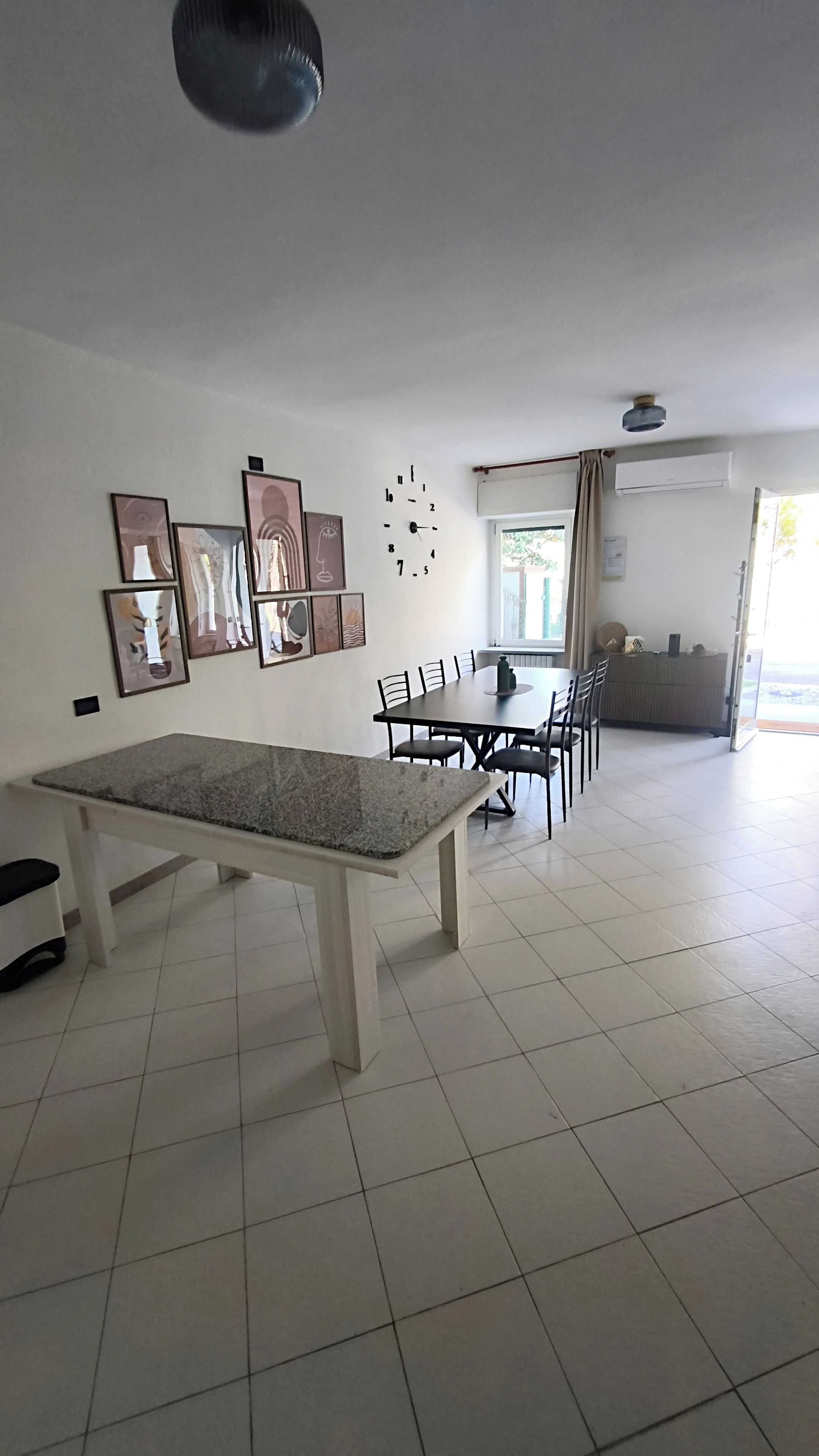 Ein geräumiges Zimmer mit Esstisch, Stühlen, einer Uhr und gerahmten Kunstwerken an der Wand.