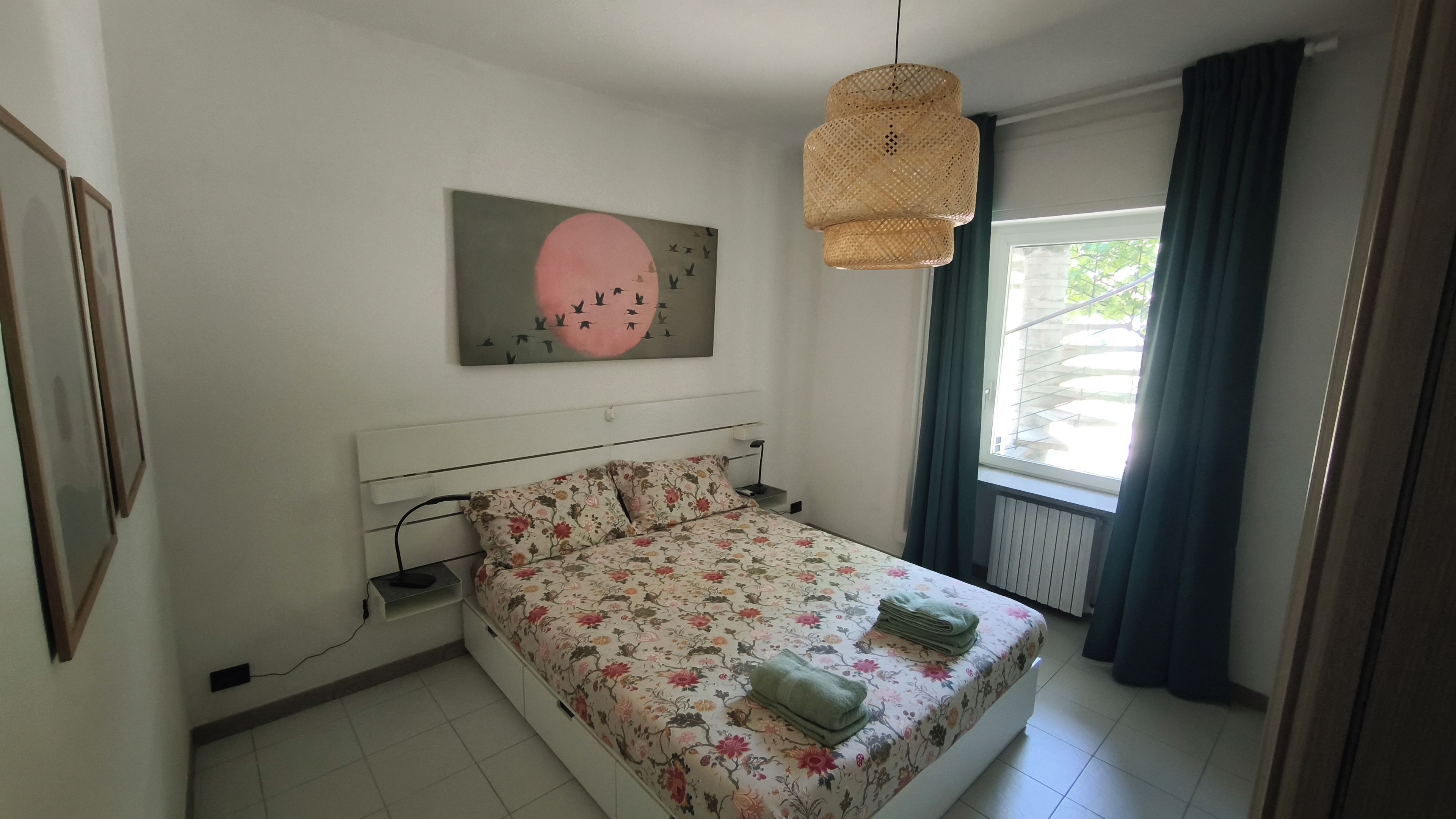 Un dormitorio con una cama, una lámpara colgante, una ventana con cortinas y un cuadro en la pared.