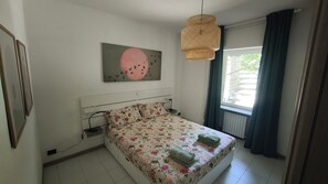 A bedroom with a bed, a hanging light fixture, a window with curtains, and a painting on the wall.