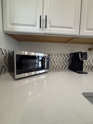 Una cocina con armarios blancos, un microondas de acero inoxidable y una cafetera sobre una encimera blanca.