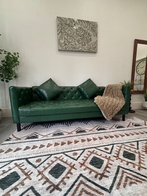 Un sofá de cuero verde con una manta, una alfombra estampada, una obra de arte abstracta colgada en la pared y un espejo con reloj.