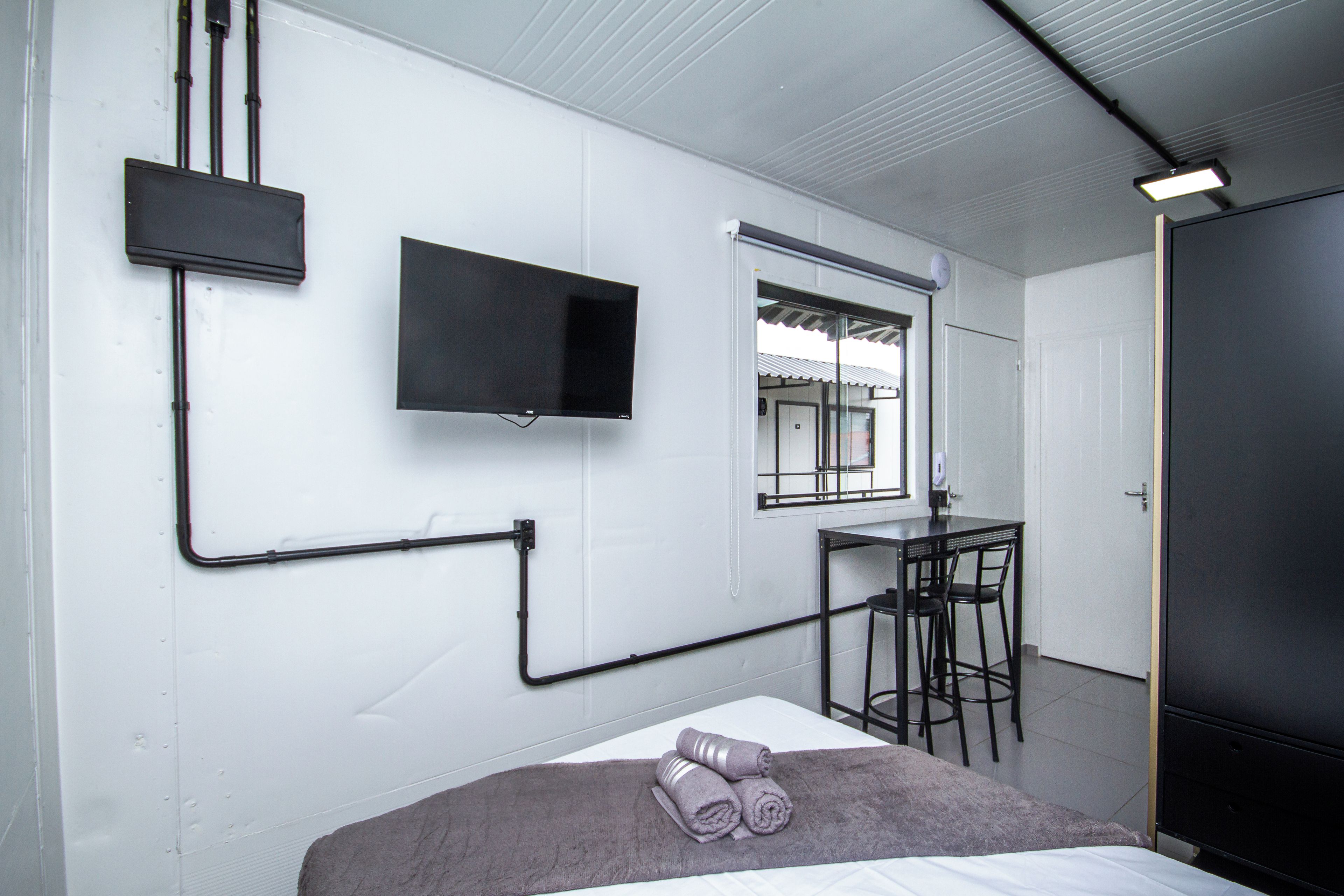 Quarto com cama, televisão de tela plana na parede, janela e pequena mesa com cadeiras.