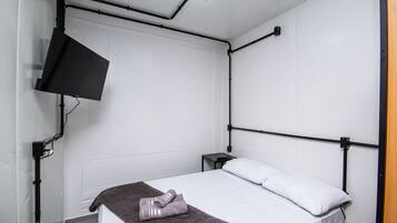 A single bed with a white mattress and pillows, a dark brown blanket, a remote control on a small bedside table, and a wall-mounted TV.