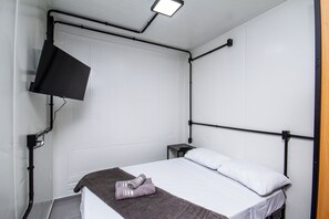 Una cama individual con colchón y almohadas blancas, una manta marrón oscuro, un mando a distancia sobre una mesita de noche pequeña y un televisor montado en la pared.