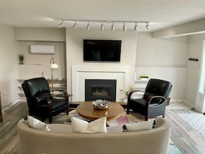 A living room with a fireplace, two leather armchairs, a sofa, a coffee table, and a flat-screen TV mounted above the fireplace.