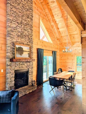 A spacious room with a stone fireplace, wooden ceiling beams, a dining table with chairs, and a chandelier.