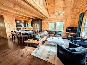 A log cabin interior with a living room, dining area, kitchen, and a large window.