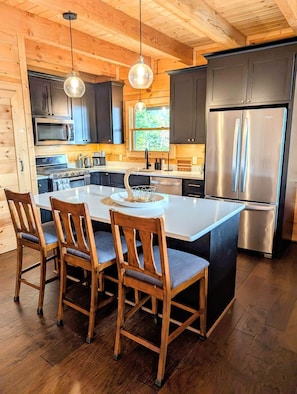 A modern kitchen with a large island, dark cabinetry, stainless steel appliances, and wooden chairs.