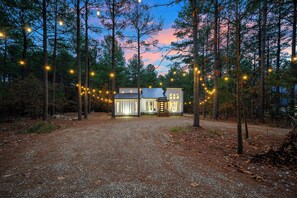 A two-story cabin with a gravel driveway, surrounded by tall trees and string lights. - Large Deck! Hot tub! HorseShoe Set! Outdoor Dining (Broken Bow)