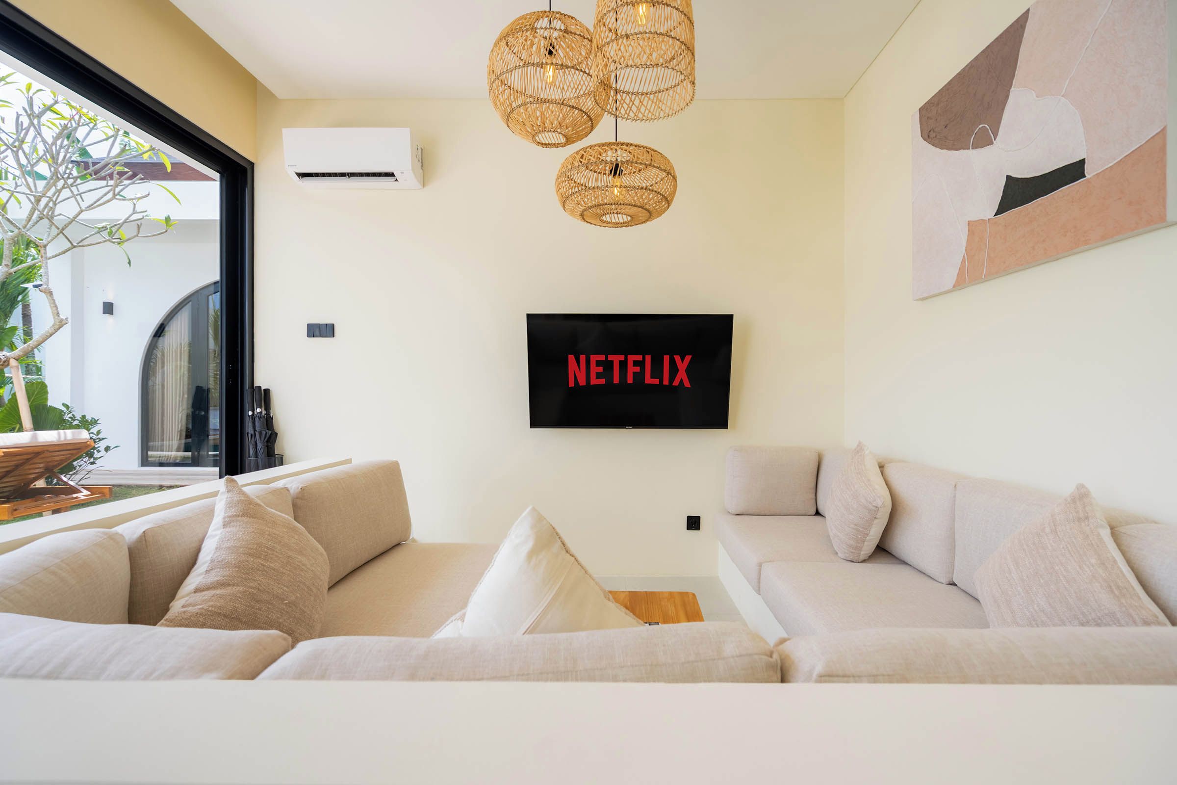 Ein modernes Wohnzimmer mit einem großen Flachbildfernseher an der Wand, einer Korbschwinge-Pendelleuchte und einem beigefarbenen Sofa mit Kissen.