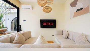 Ein modernes Wohnzimmer mit einem großen Flachbildfernseher an der Wand, einer Korbschwinge-Pendelleuchte und einem beigefarbenen Sofa mit Kissen.