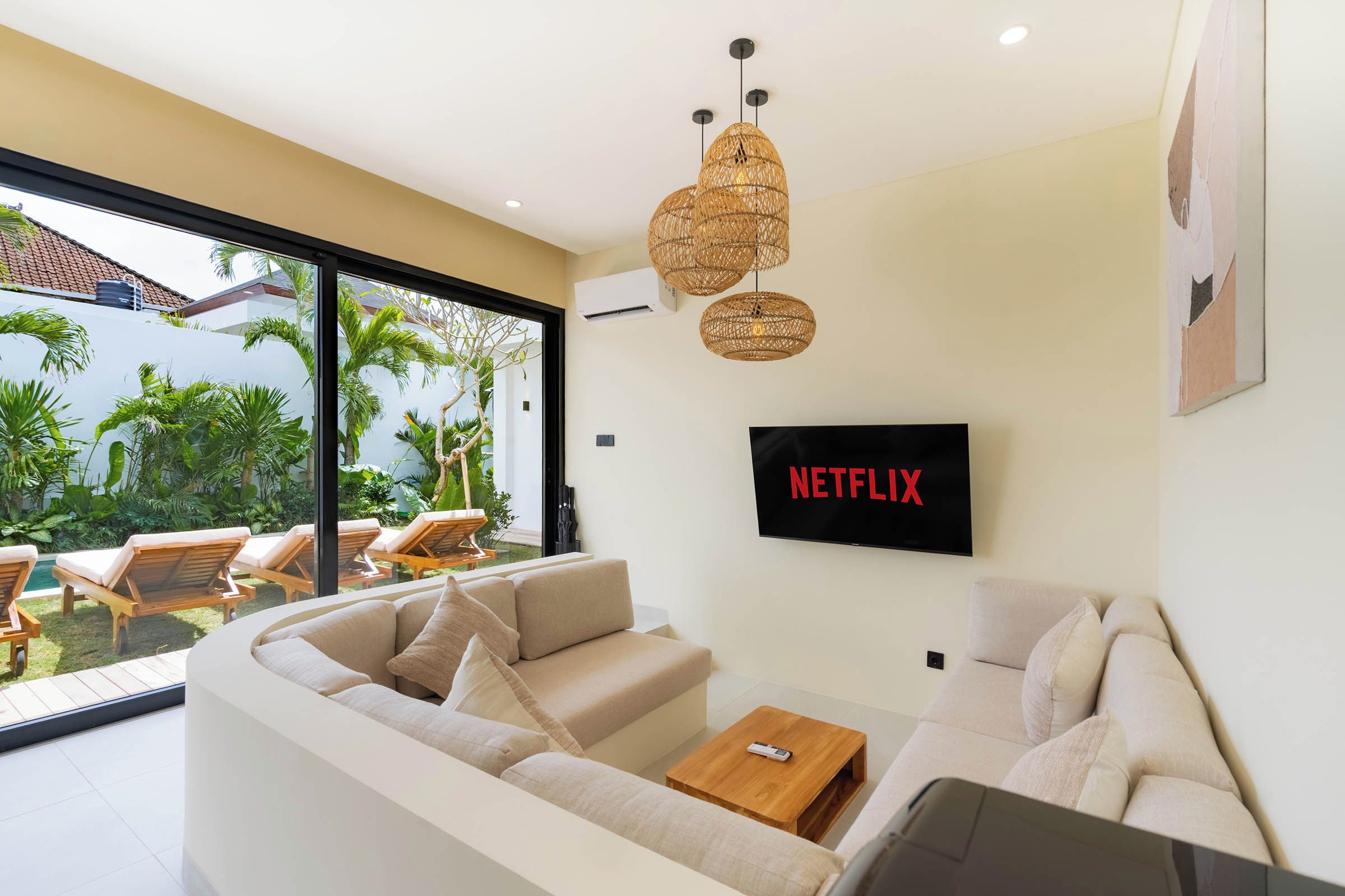 Un salon moderne avec un grand téléviseur fixé au mur, un canapé, une table basse et une vue sur un jardin grâce à de grandes fenêtres.