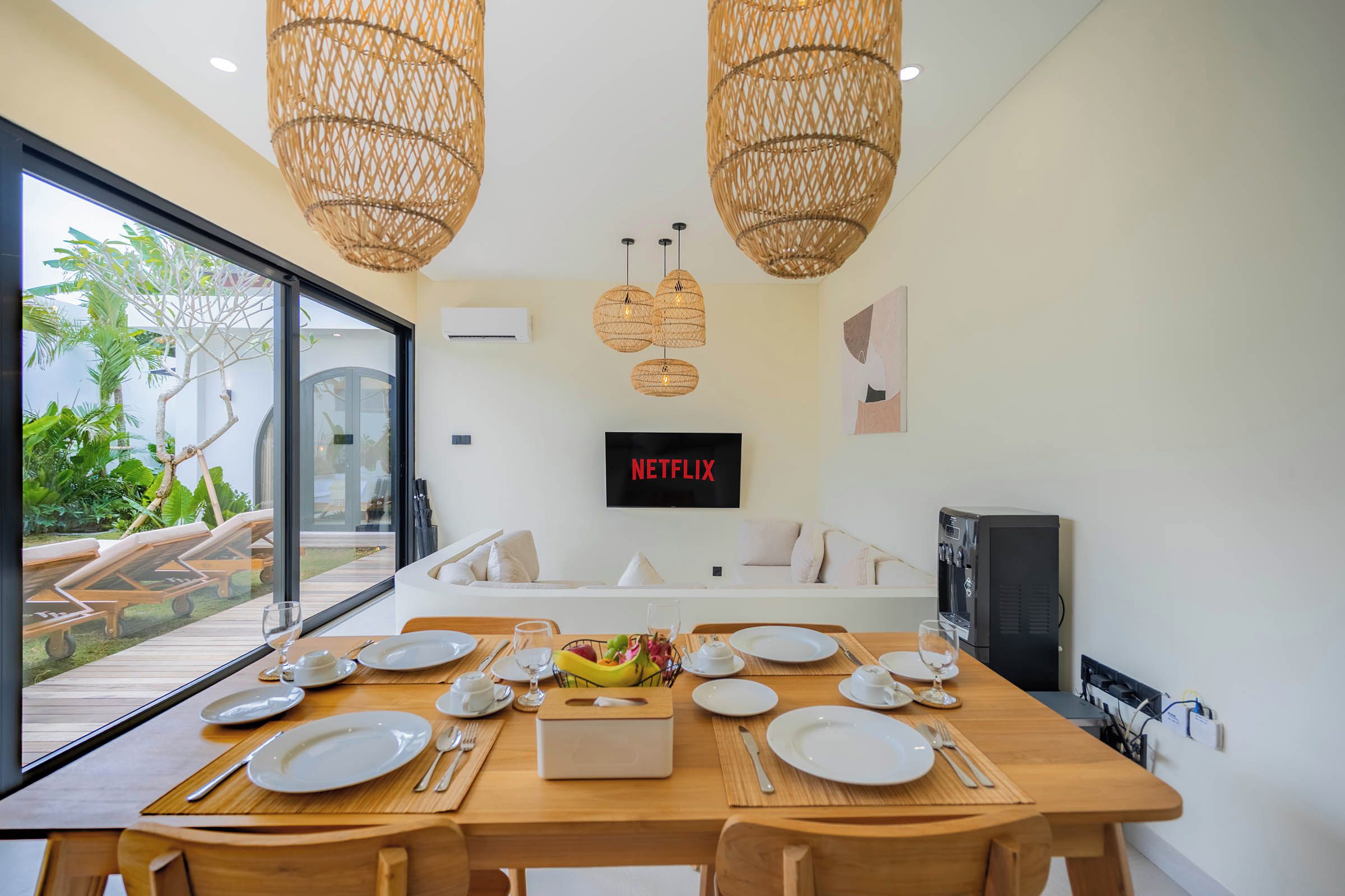 A dining area with a wooden table set for four, a TV mounted on the wall, and a view of the outdoors through large windows.