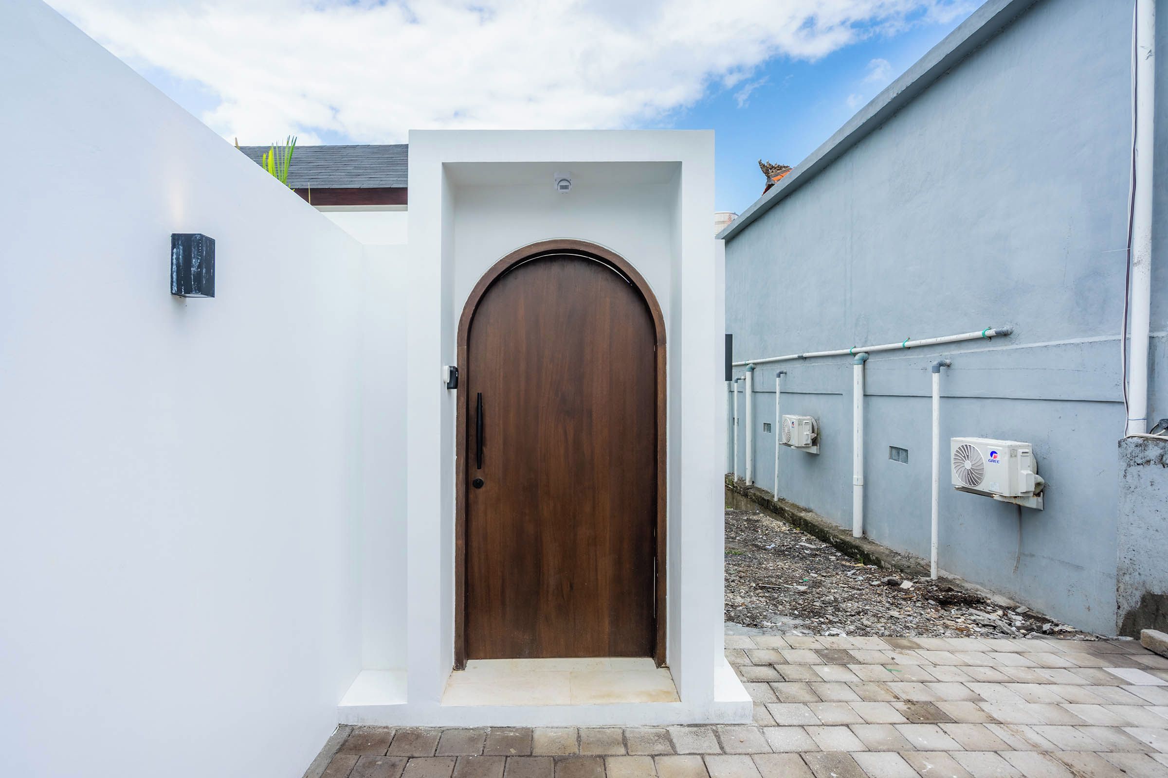 Un sendero estrecho que conduce a una puerta de madera con marco blanco, flanqueado por paredes blancas y un edificio gris con tuberías visibles y un equipo de aire acondicionado.