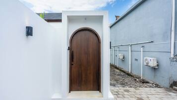 Un sendero estrecho que conduce a una puerta de madera con marco blanco, flanqueado por paredes blancas y un edificio gris con tuberías visibles y un equipo de aire acondicionado.