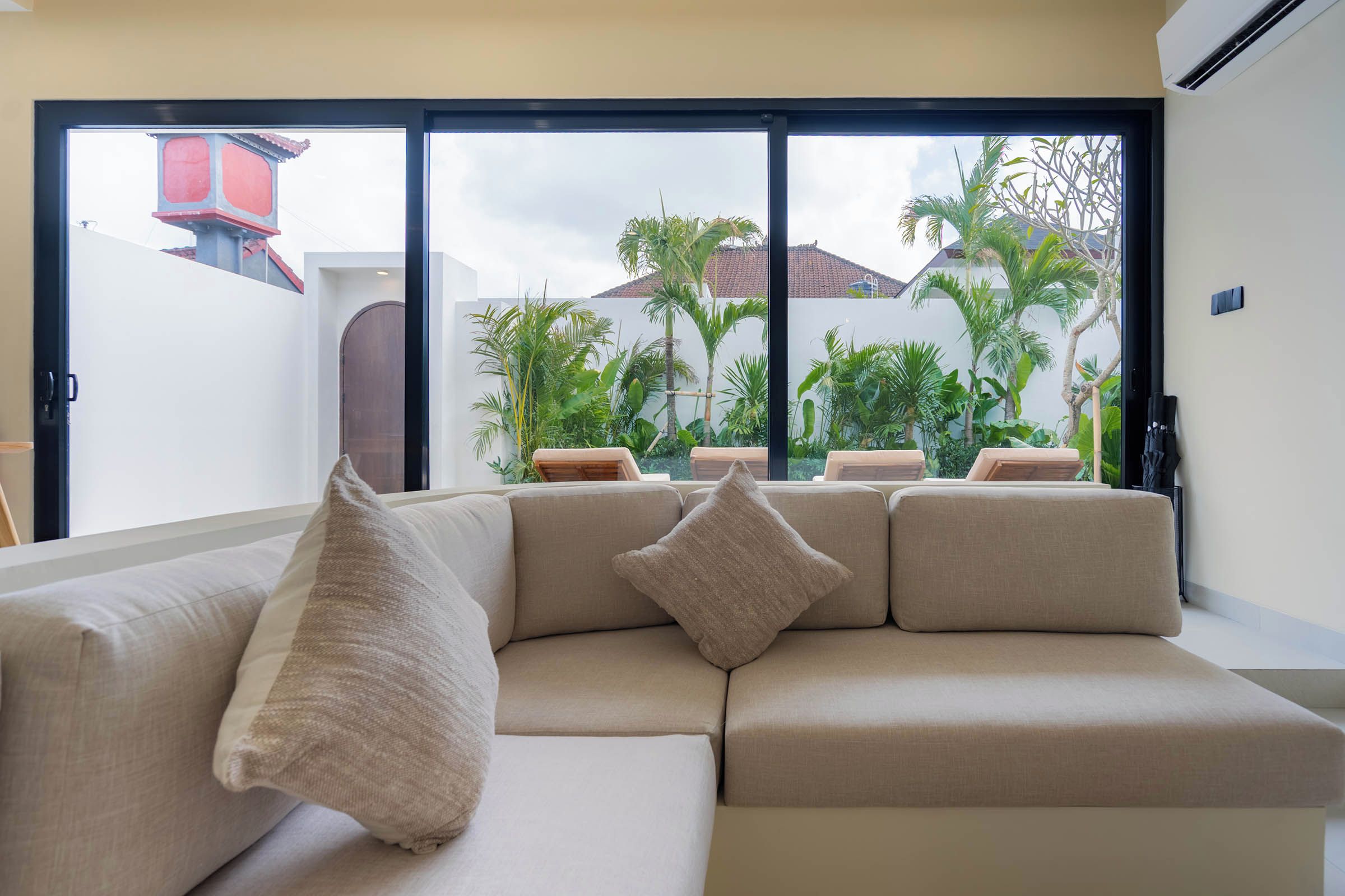 A beige sectional sofa with two pillows, a sliding glass door leading to a balcony with a view of palm trees and a building.