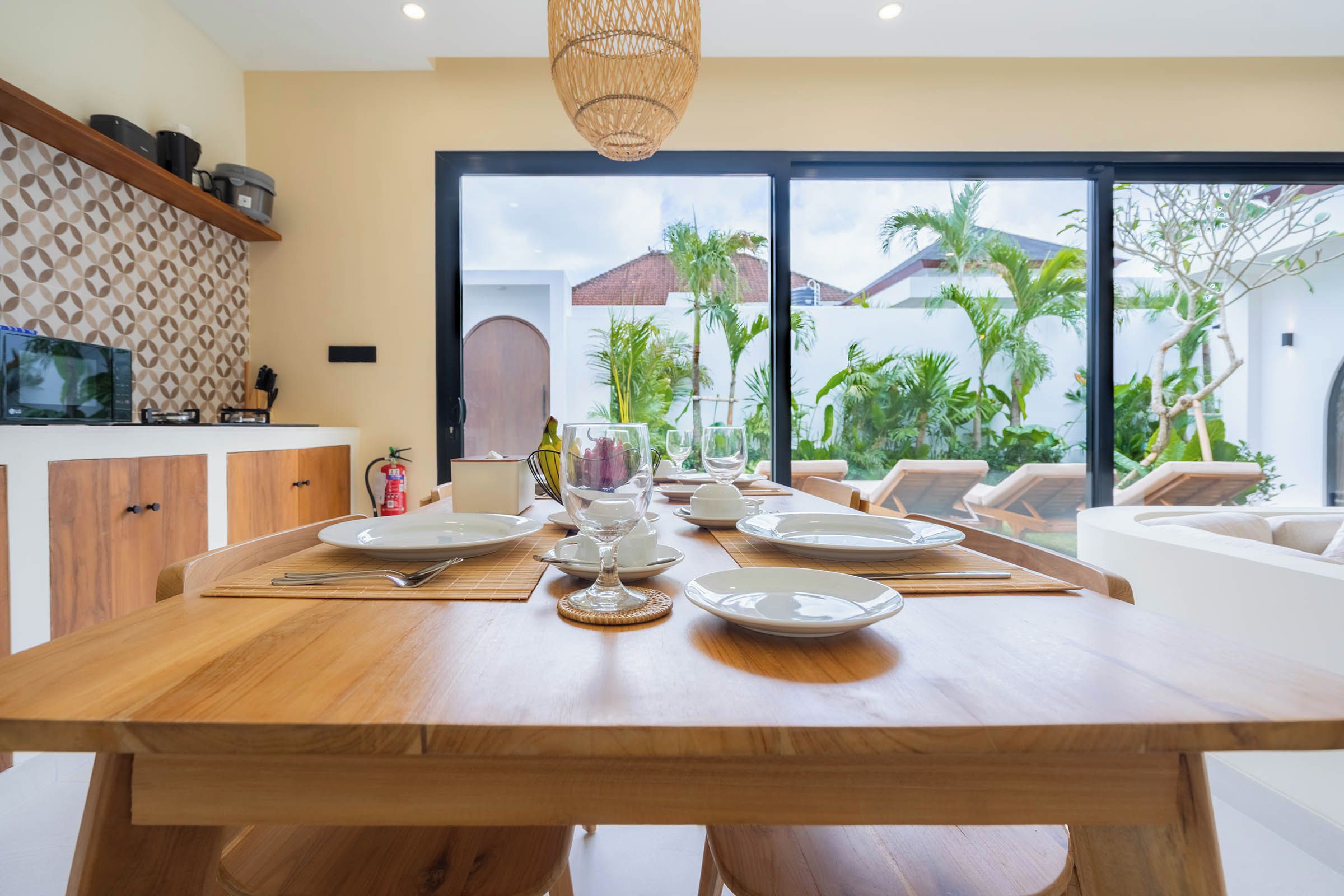 Uma sala de jantar com uma mesa de madeira posta para refeição, uma televisão à esquerda e vista para palmeiras através de grandes janelas.