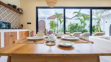 Uma sala de jantar com uma mesa de madeira posta para refeição, uma televisão à esquerda e vista para palmeiras através de grandes janelas.