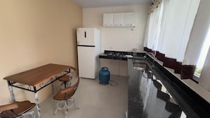 A compact kitchen with a wooden table and chairs, a white refrigerator, a gas stove, and a blue gas cylinder. - MANDACARU POUSADA (Aracruz)