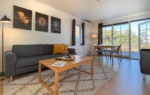 A modern living room with a grey sofa, a wooden coffee table, and a dining area with a table and chairs. There are framed pictures on the wall, and the room has large windows. - 2 bedroom cozy home in Læsø (Læsø)