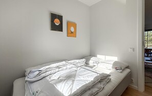 Una cama individual con ropa de cama y almohadas blancas, dos cuadros en la pared y un ventanal que deja entrar luz natural.