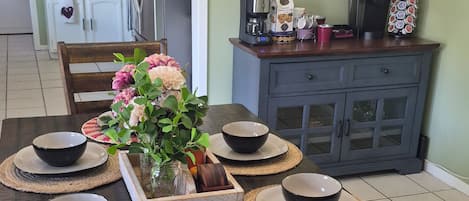 A dining table set with black bowls and plates, surrounded by woven placemats, in a room with a chandelier, kitchen cabinets, and a refrigerator.