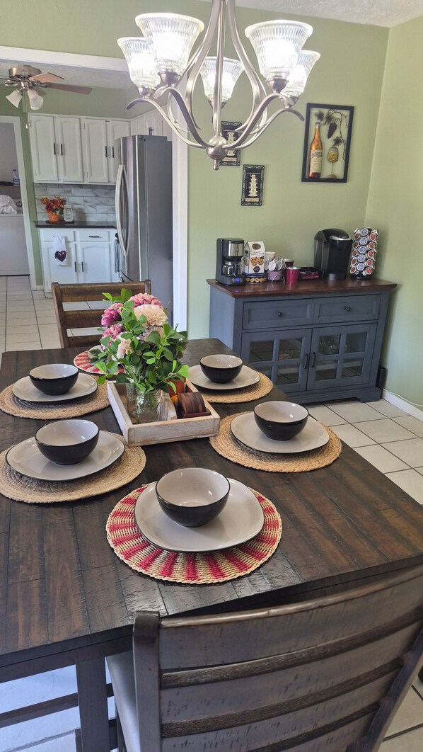 A dining table set with black bowls and plates, surrounded by woven placemats, in a room with a chandelier, kitchen cabinets, and a refrigerator.