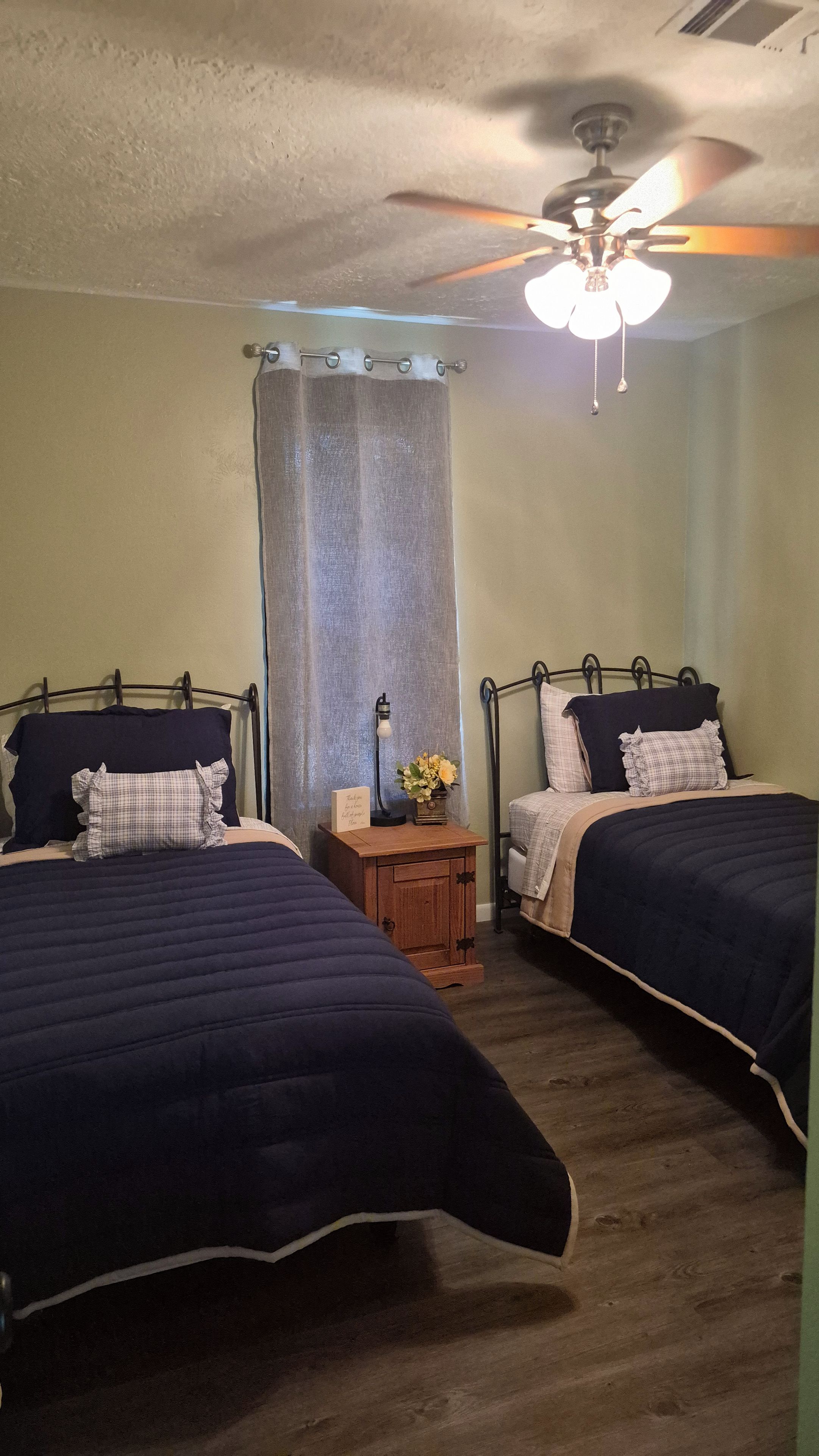 Une chambre avec deux lits, un ventilateur de plafond, une table de chevet avec une lampe et un rideau.
