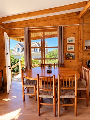 A wooden interior with a round dining table surrounded by chairs, a window with curtains, and framed pictures on the wall.
