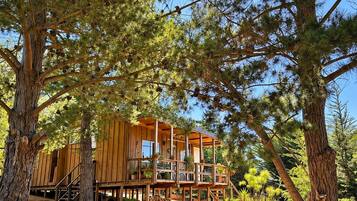 Una cabaña de madera con varios niveles, rodeada de árboles y suelo de tierra.