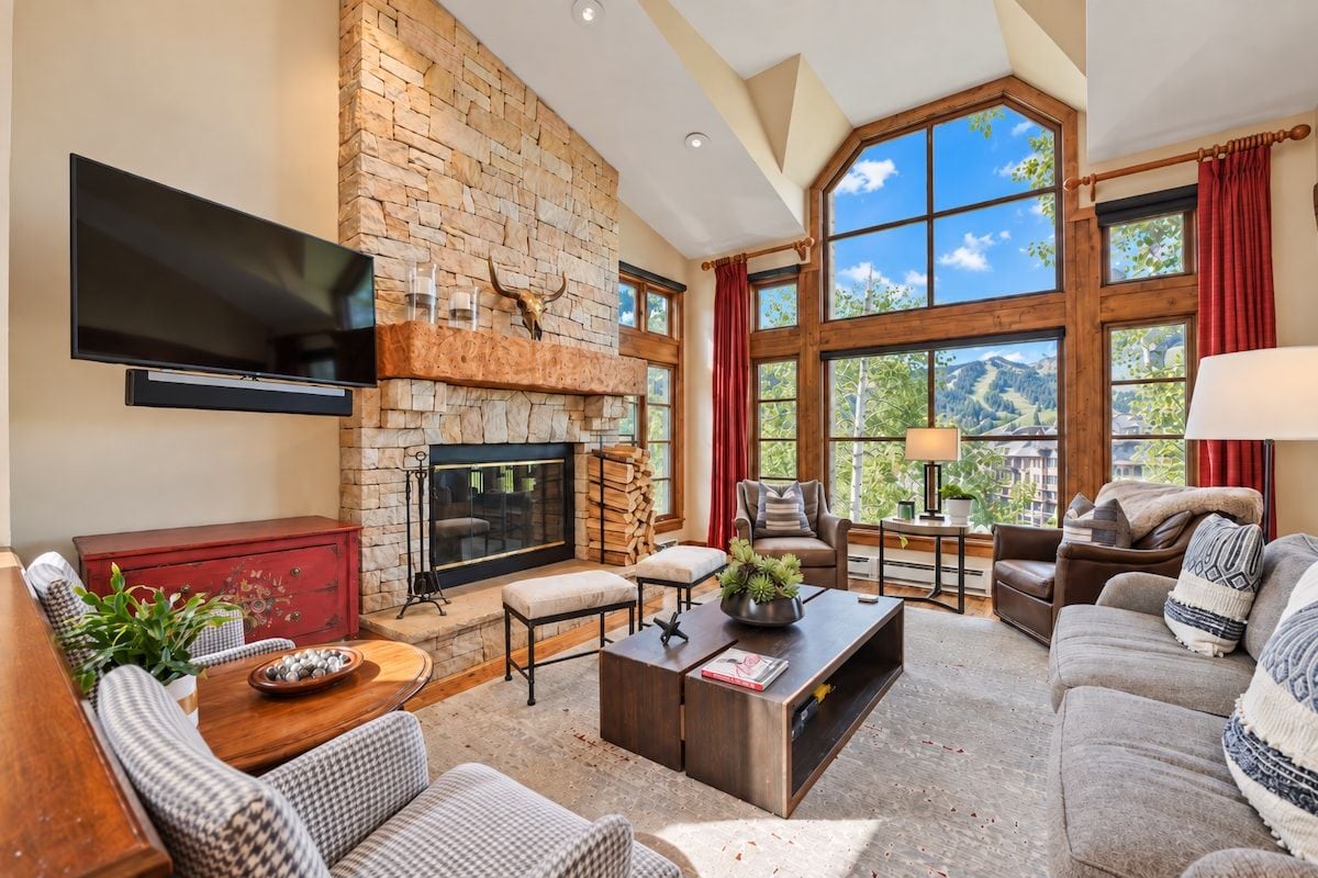 A spacious living room with a stone fireplace, large windows, and a flat-screen TV mounted on the wall.