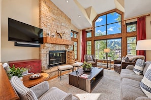 A spacious living room with a stone fireplace, large windows, and a flat-screen TV mounted on the wall. - Beaver Creek True Ski-In/Ski-Out | Private Hot Tub (Beaver Creek)