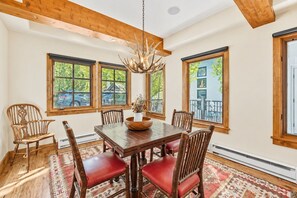 A dining area with a wooden table, red cushioned chairs, a chandelier, and large windows with views of greenery and buildings. - Beaver Creek True Ski-In/Ski-Out | Private Hot Tub (Beaver Creek)