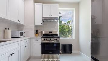 Cozinha com armários brancos, fogão, micro-ondas e janela com vista para árvores.