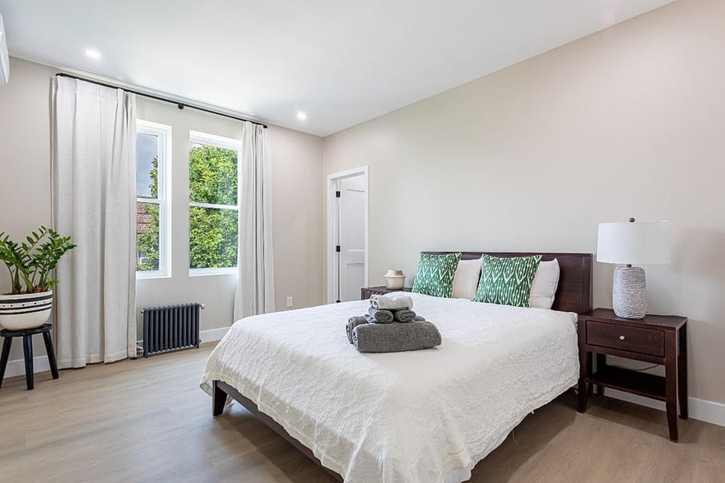 Quarto com cama grande, mesa de cabeceira, luminária, planta em vaso e janela com cortinas.